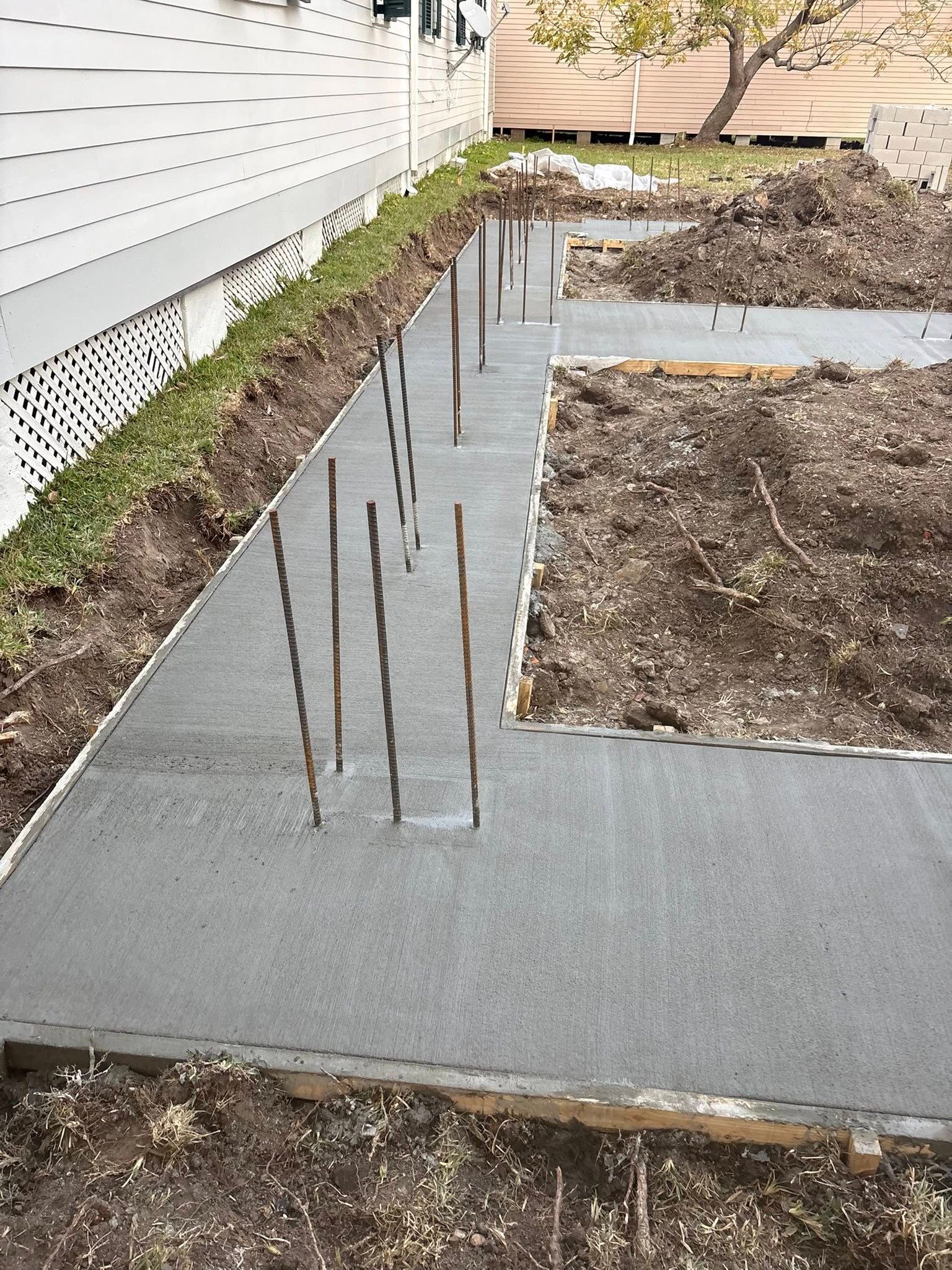 A newly poured concrete foundation with vertical steel rebar protruding from it, running alongside a house exterior.