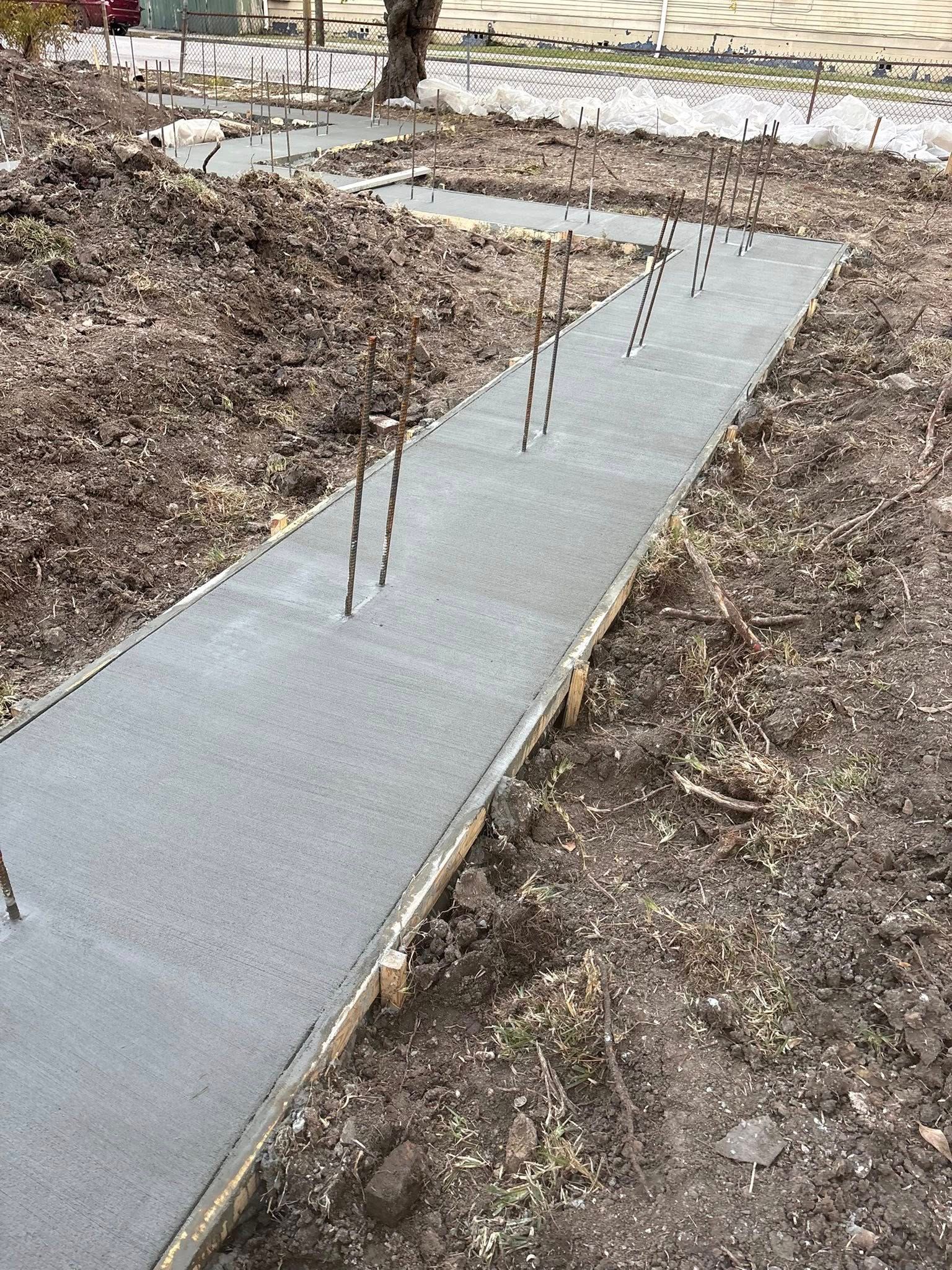 A newly poured concrete foundation footer with vertical metal rebar sticking out, set in an outdoor dirt construction site.