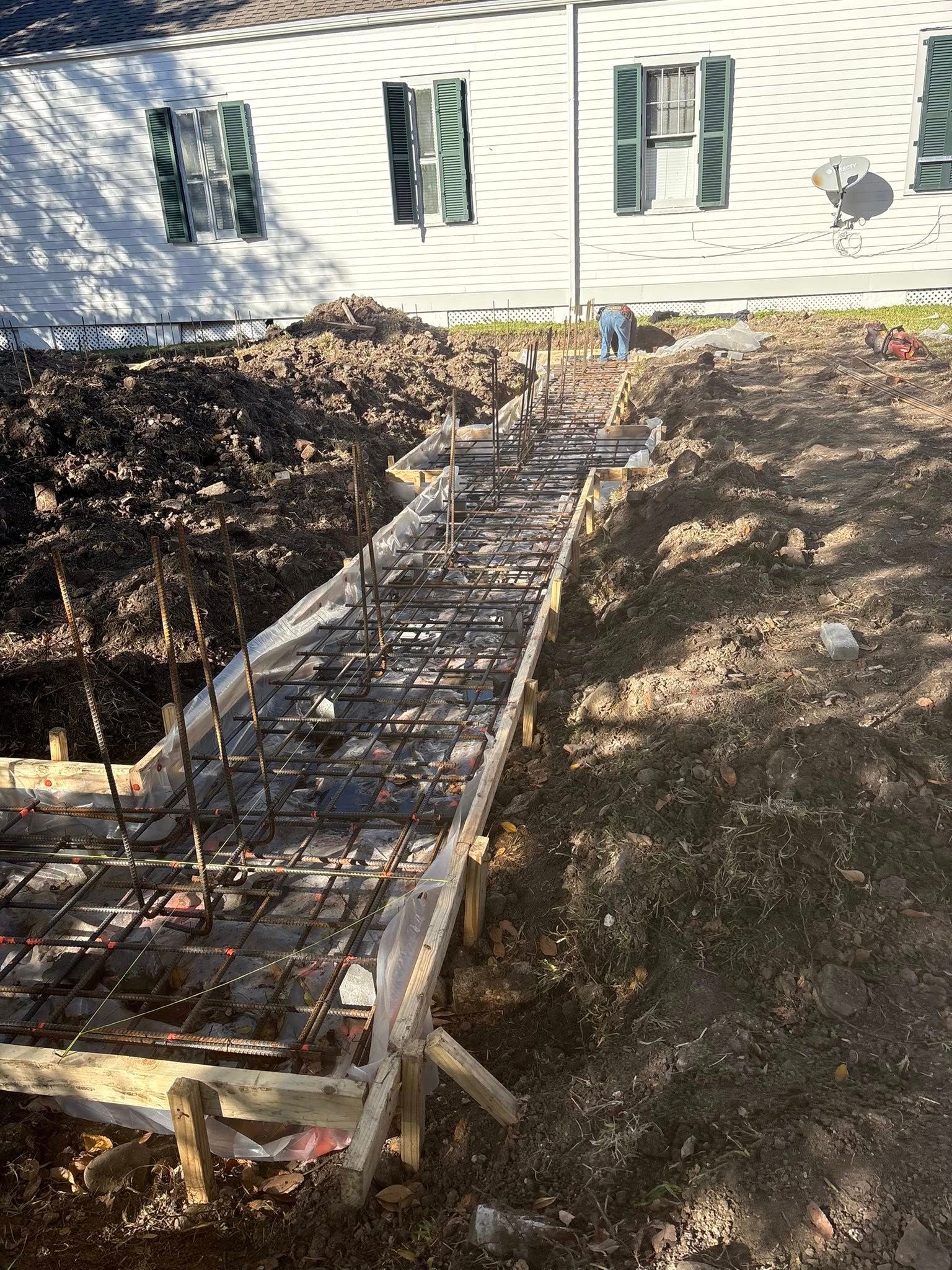 A long, narrow construction trench with wooden forms and metal rebar, positioned in front of a white house with shutters.