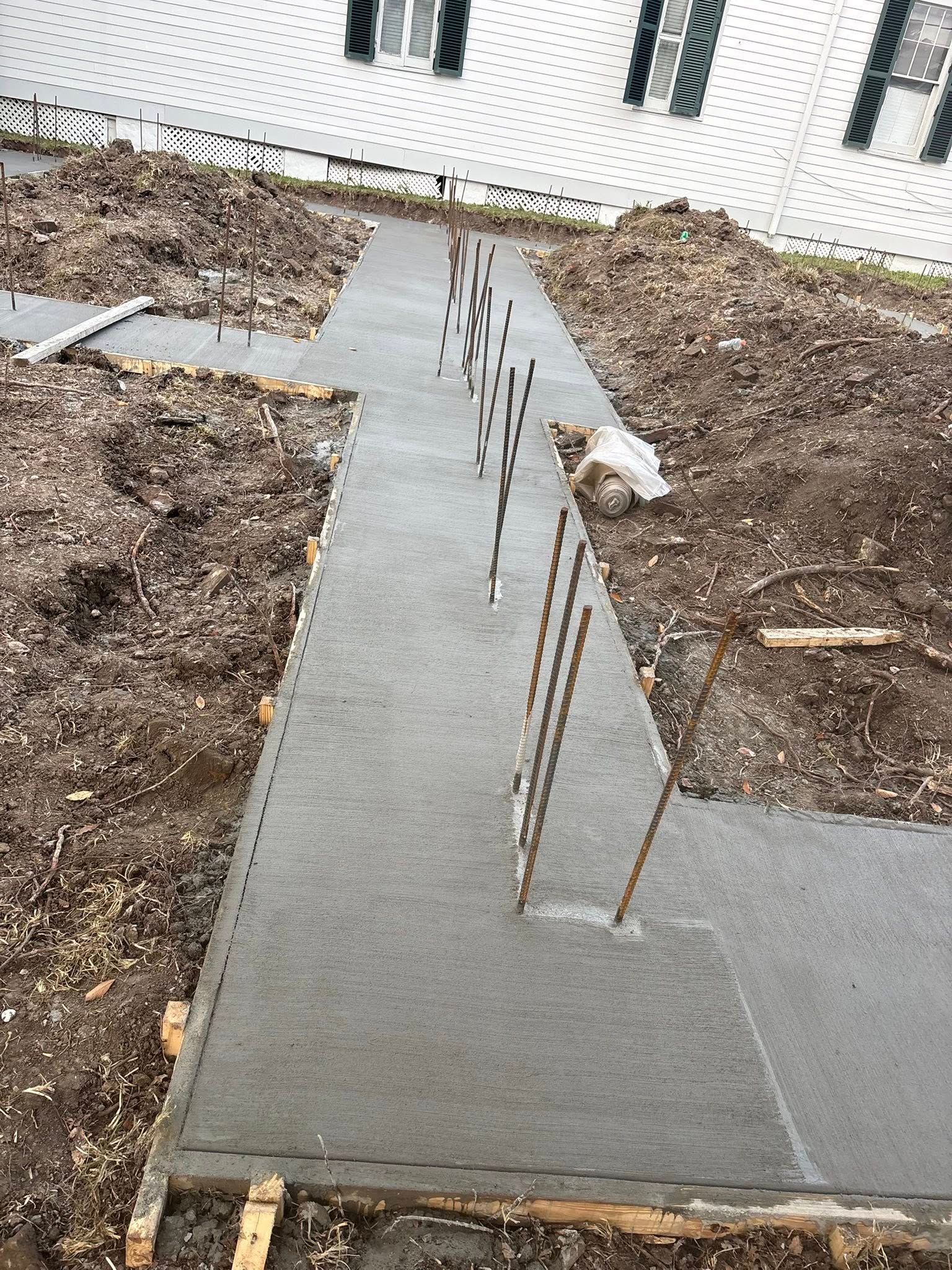 A fresh concrete foundation footing with vertical metal rebar protruding, set in an excavated dirt trench.