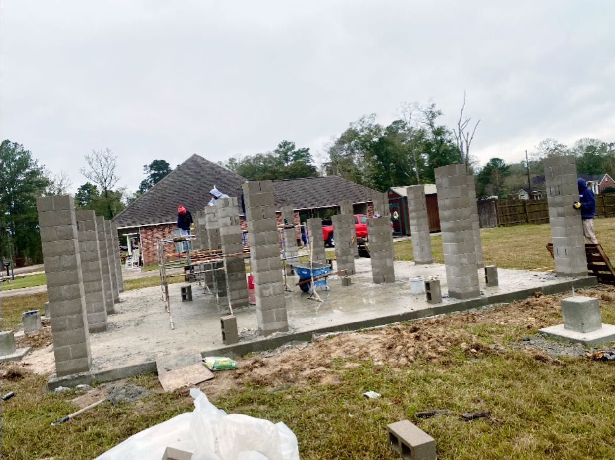 Construction workers build a structure with concrete block pillars on a concrete foundation in an outdoor setting.