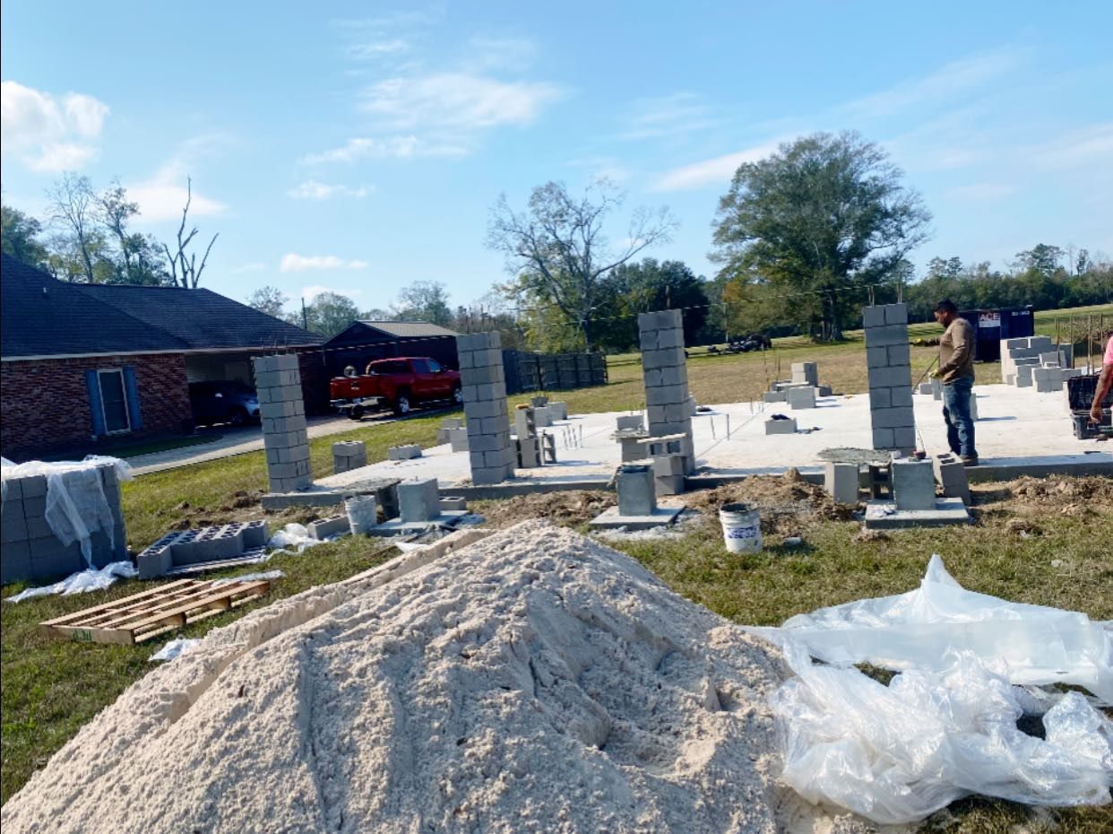 A construction site shows concrete block pillars being built on a foundation, with a pile of sand in the foreground.