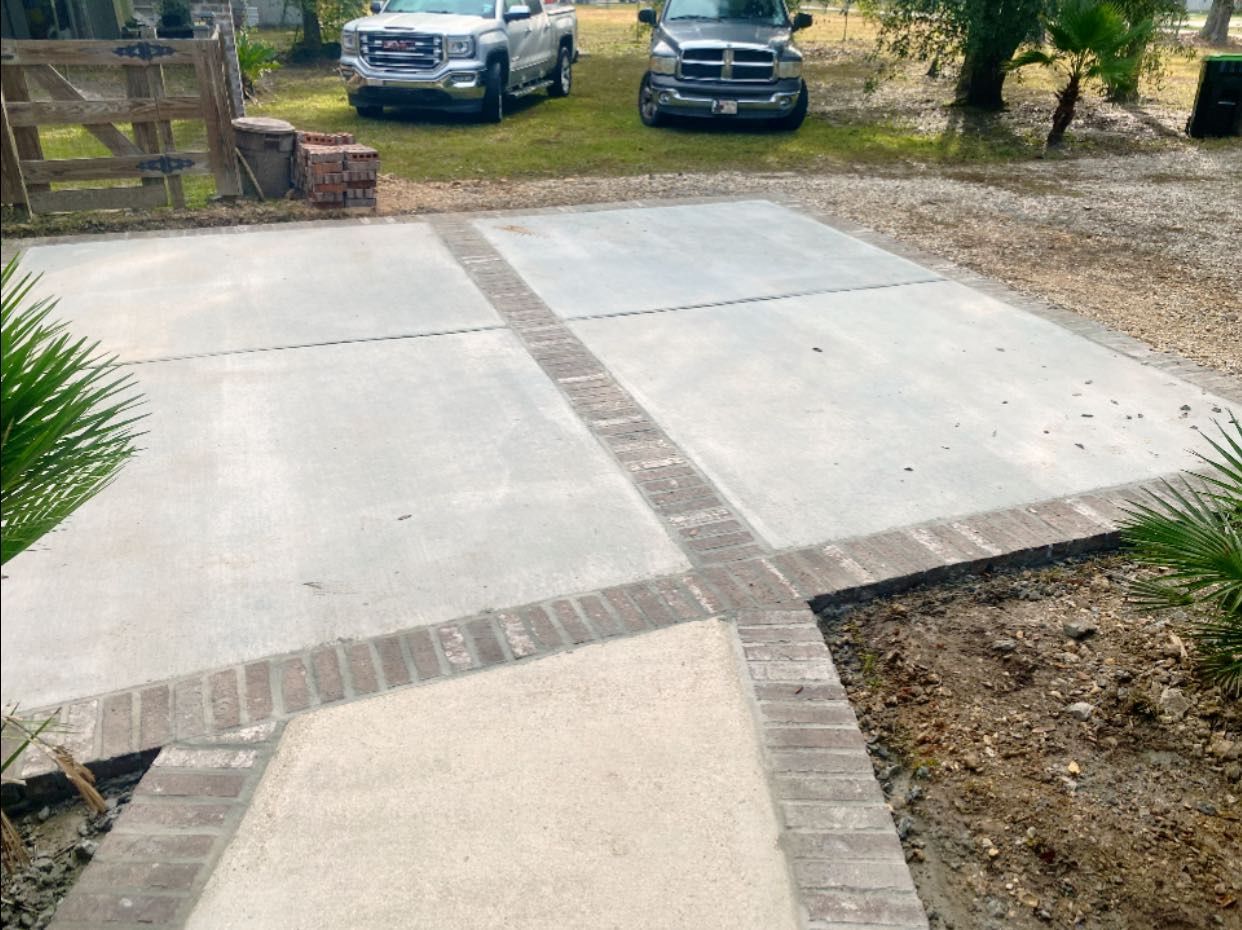 A concrete patio bordered by brick, featuring a walkway leading to a driveway with two parked pickup trucks.