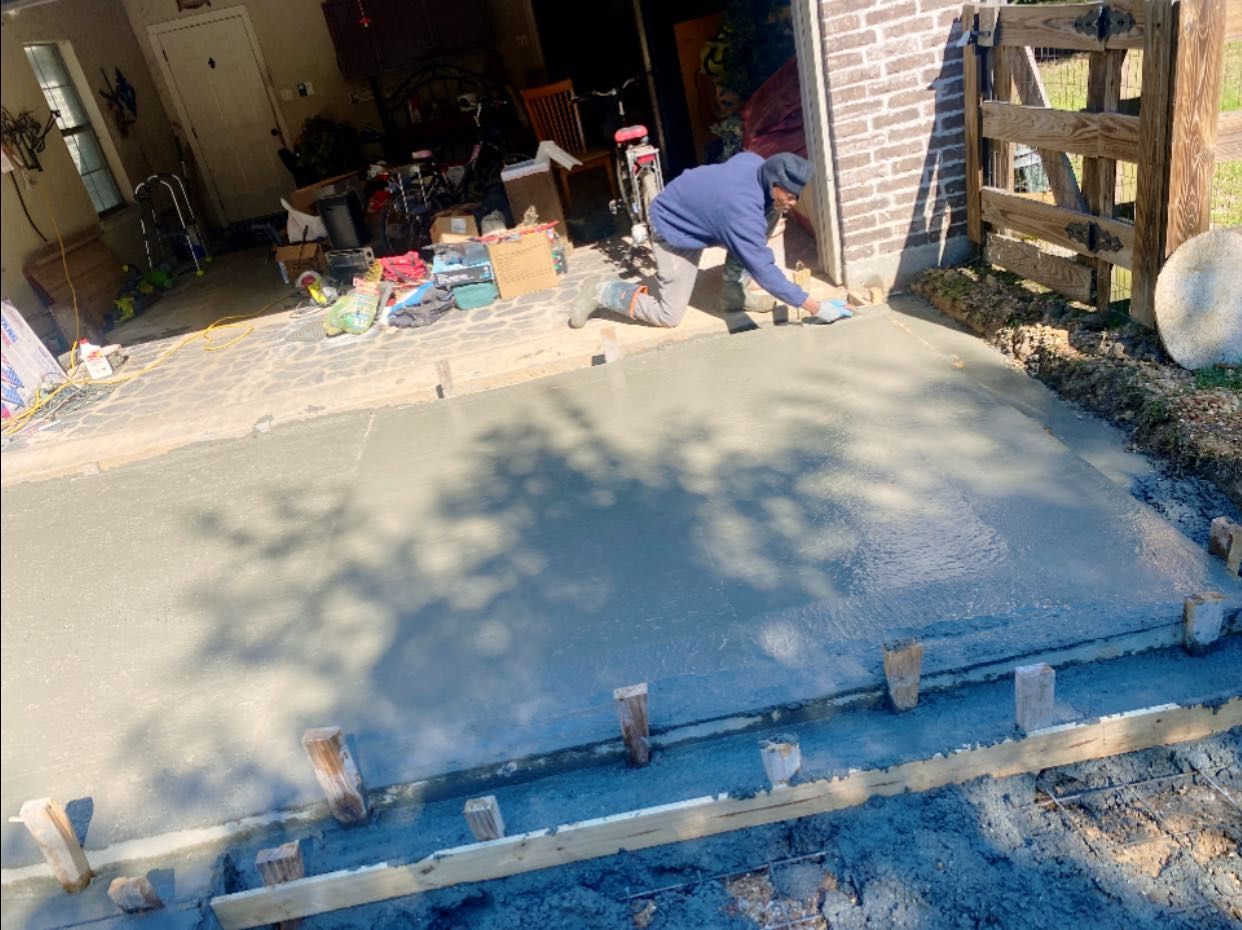 A person in a blue hoodie kneels on freshly poured concrete, smoothing the surface with a tool near a wooden garage frame.