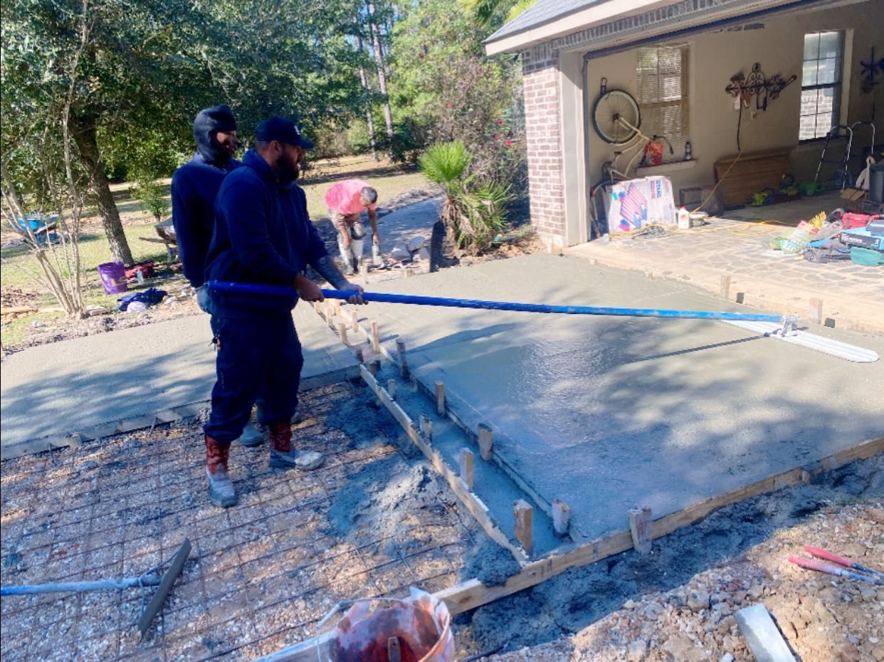 Two workers use a long-handled concrete tool to smooth freshly poured concrete in a residential driveway.