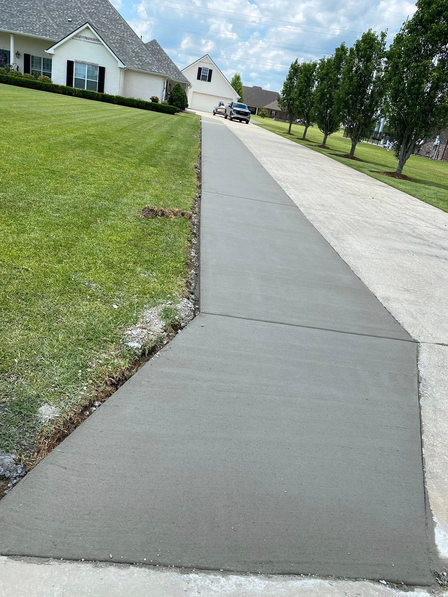 A newly poured, smooth gray concrete sidewalk runs alongside a green grassy lawn and an older paved driveway.