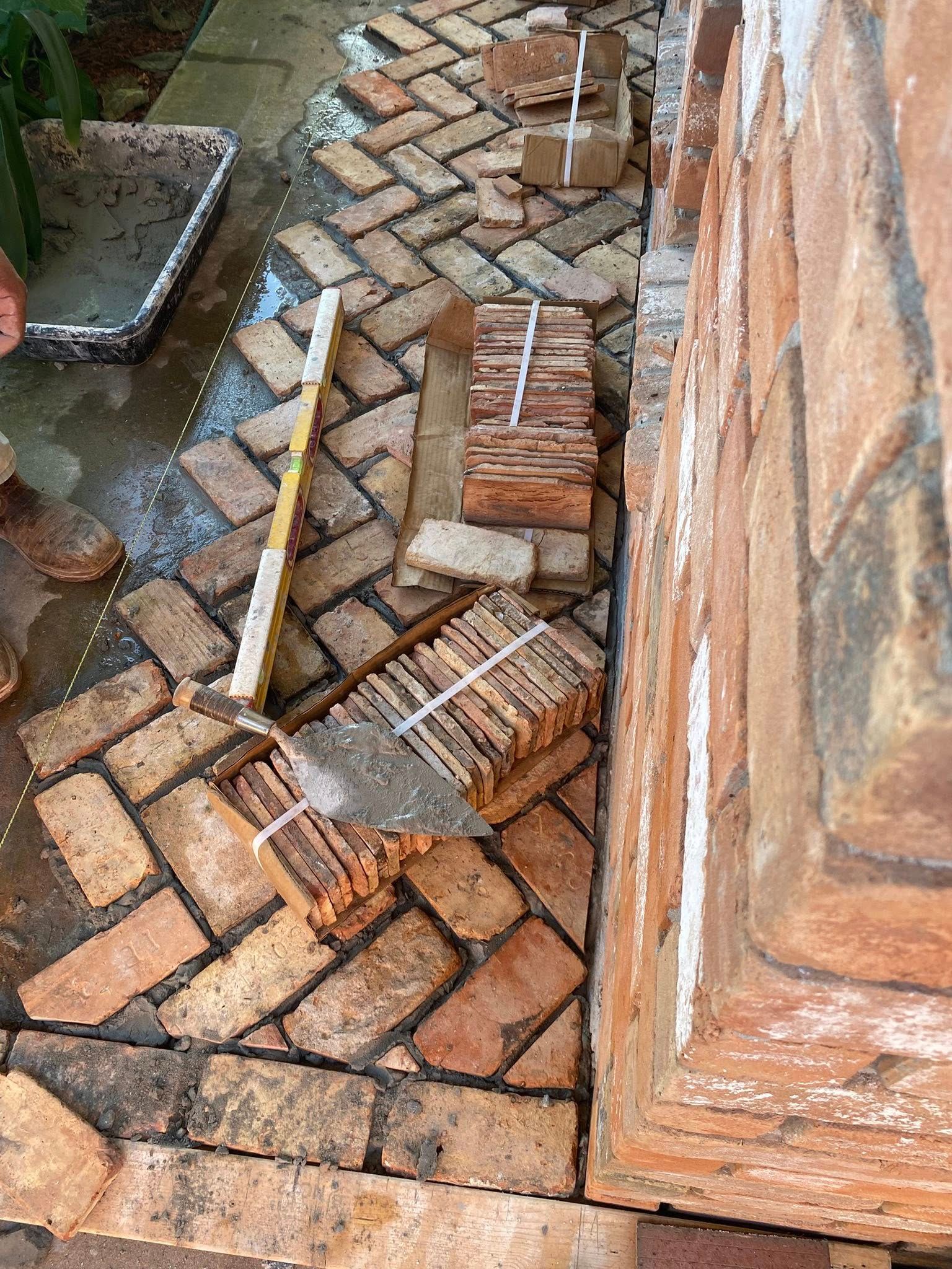 Bricks arranged in a herringbone pattern on a patio next to a brick wall, with stacks of bricks and a trowel nearby.