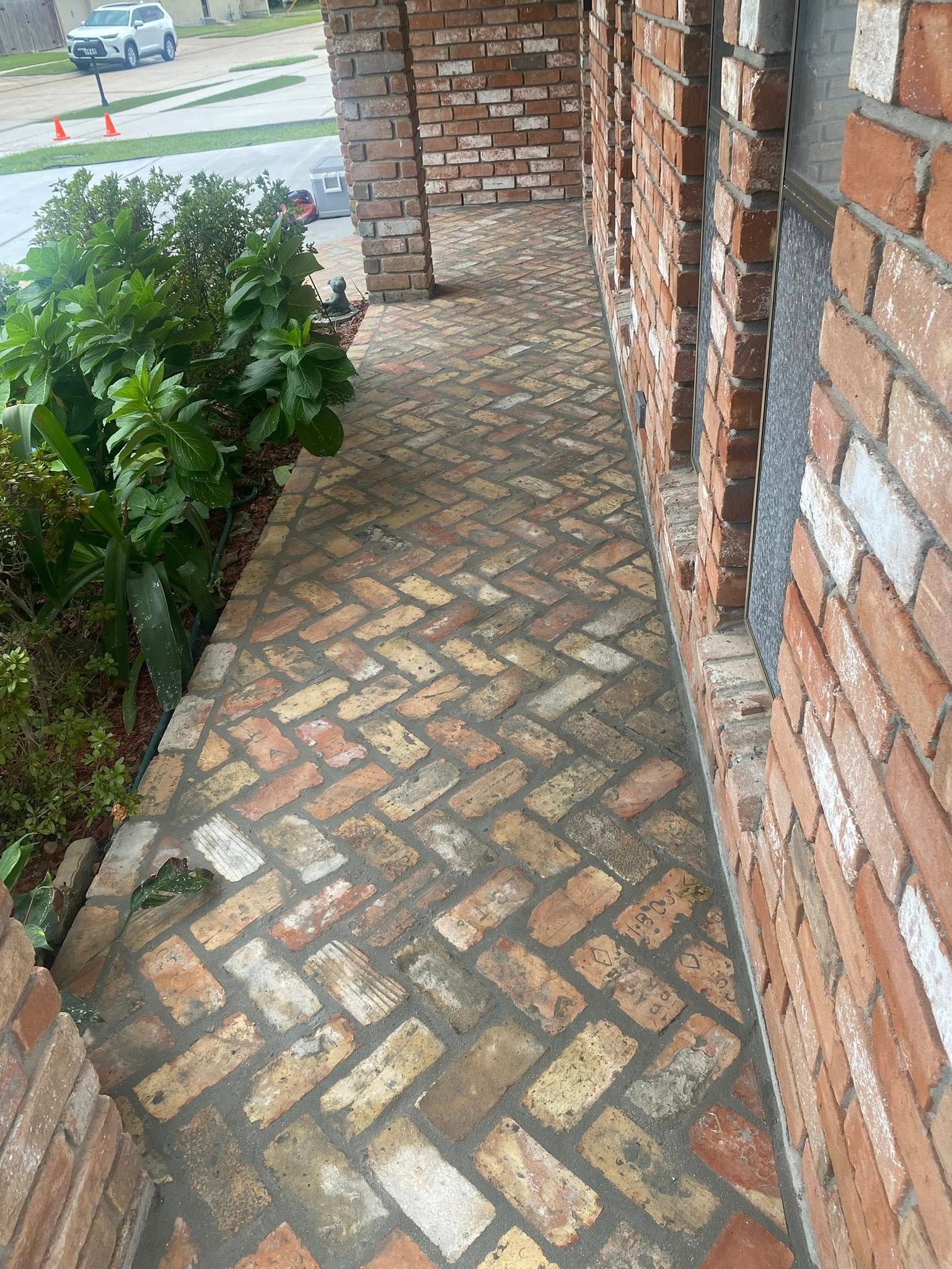 A herringbone-patterned brick walkway leads alongside a brick building and a lush green bush.