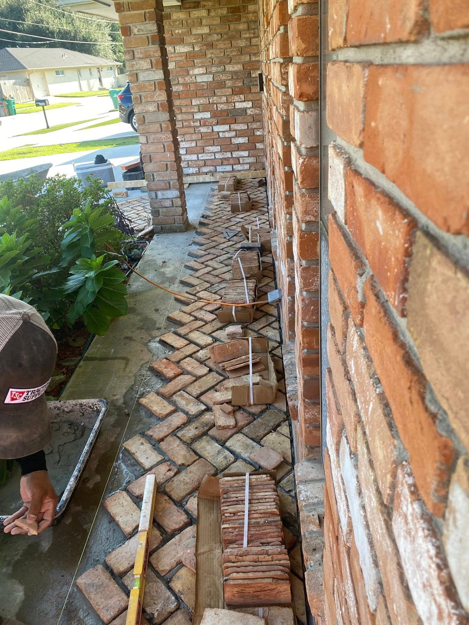 A person installing herringbone-patterned brick pavers on a walkway along a brick house exterior.