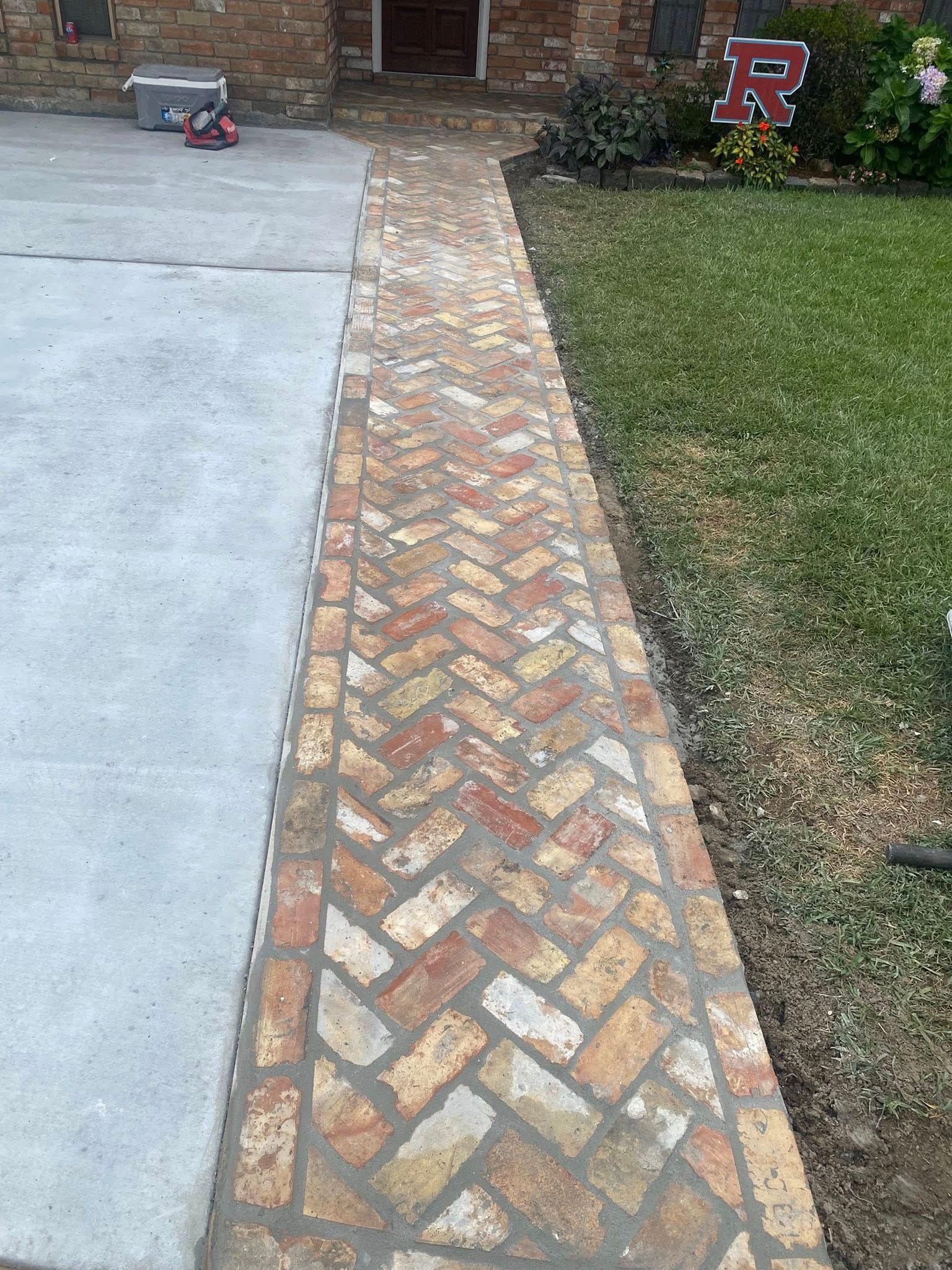 A herringbone brick walkway leads from a concrete driveway to the front entrance of a brick house.