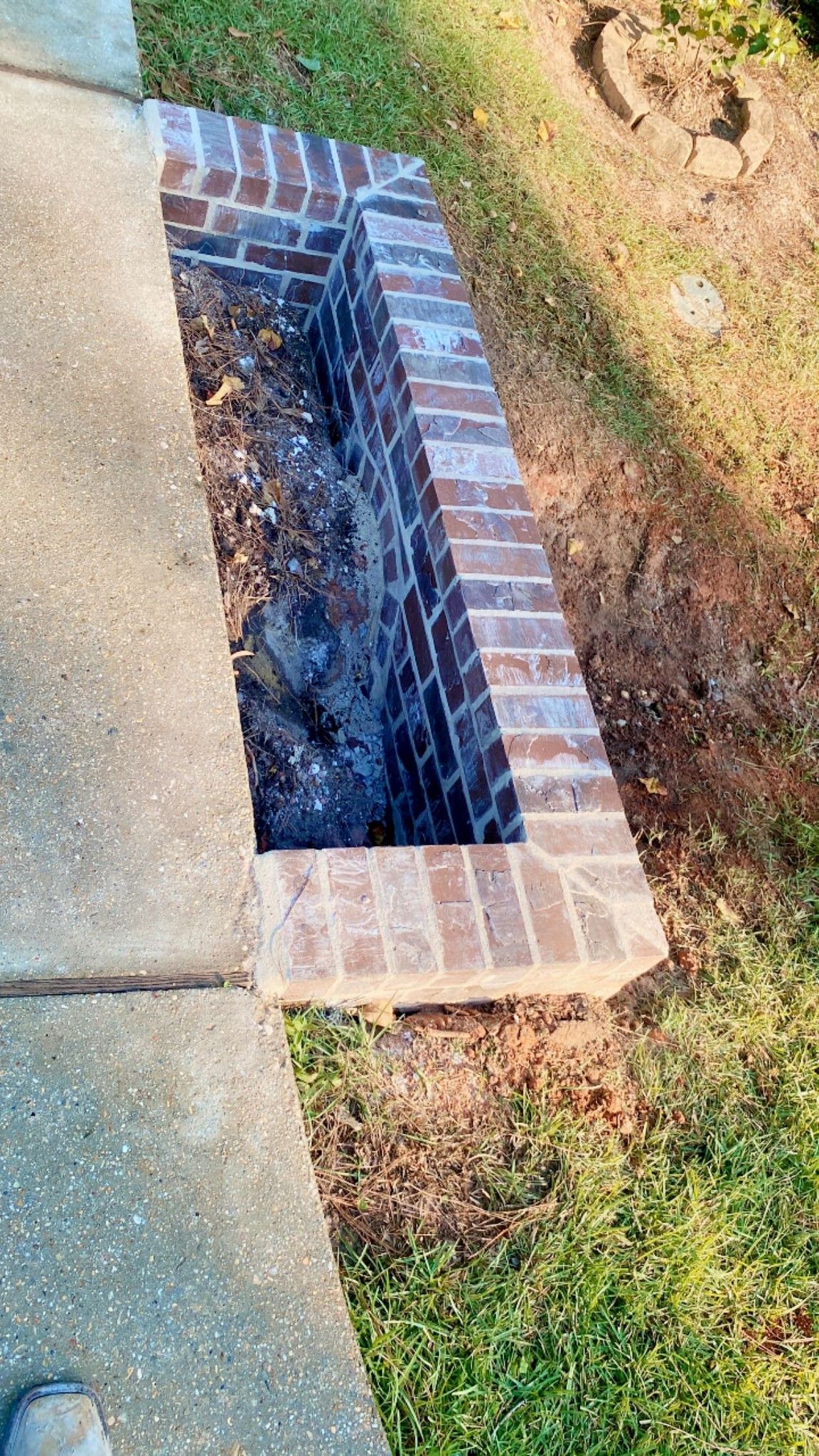 A rectangular brick planter embedded in the ground next to a concrete walkway, filled with dark soil and fallen leaves.