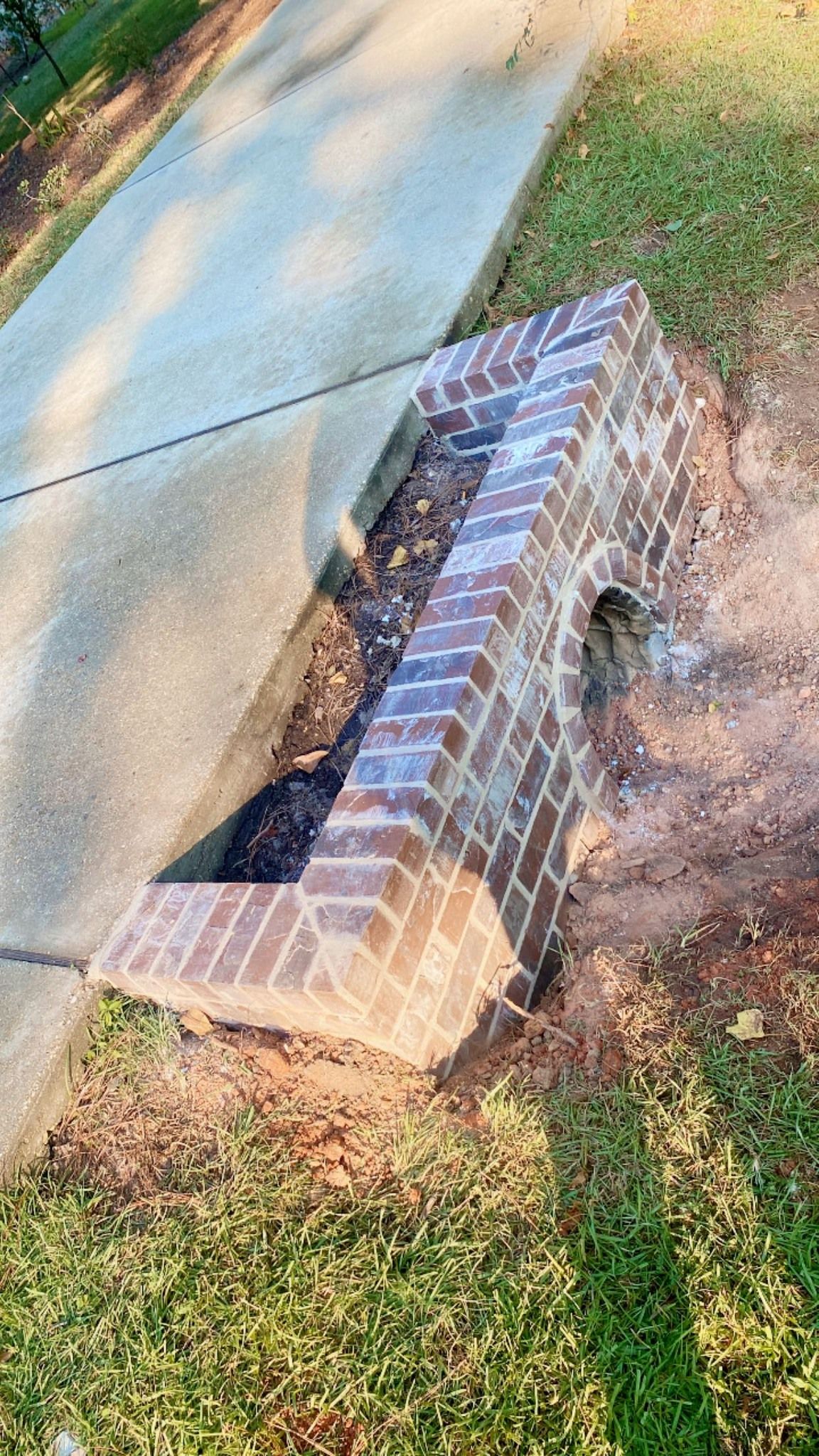 A brick drainage headwall structure sits on the edge of a concrete driveway, surrounded by grass and dirt.
