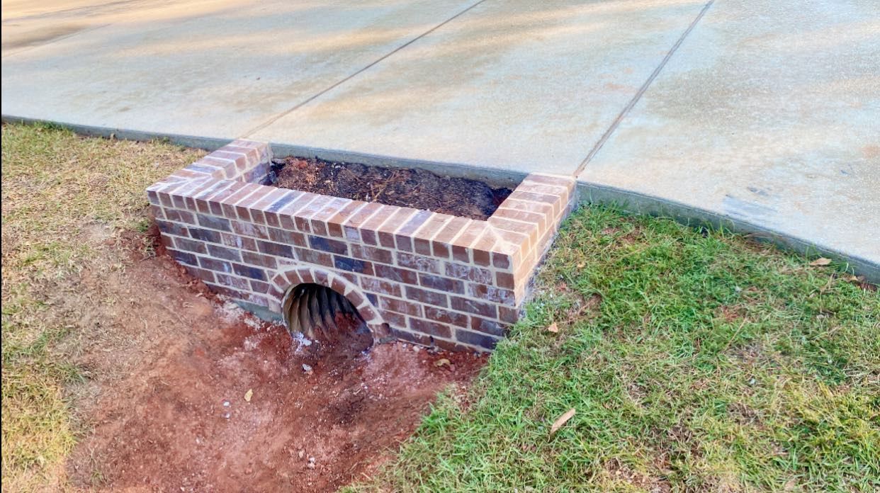 A brick drainage headwall with a corrugated metal pipe, situated at the edge of a concrete driveway and grassy lawn.