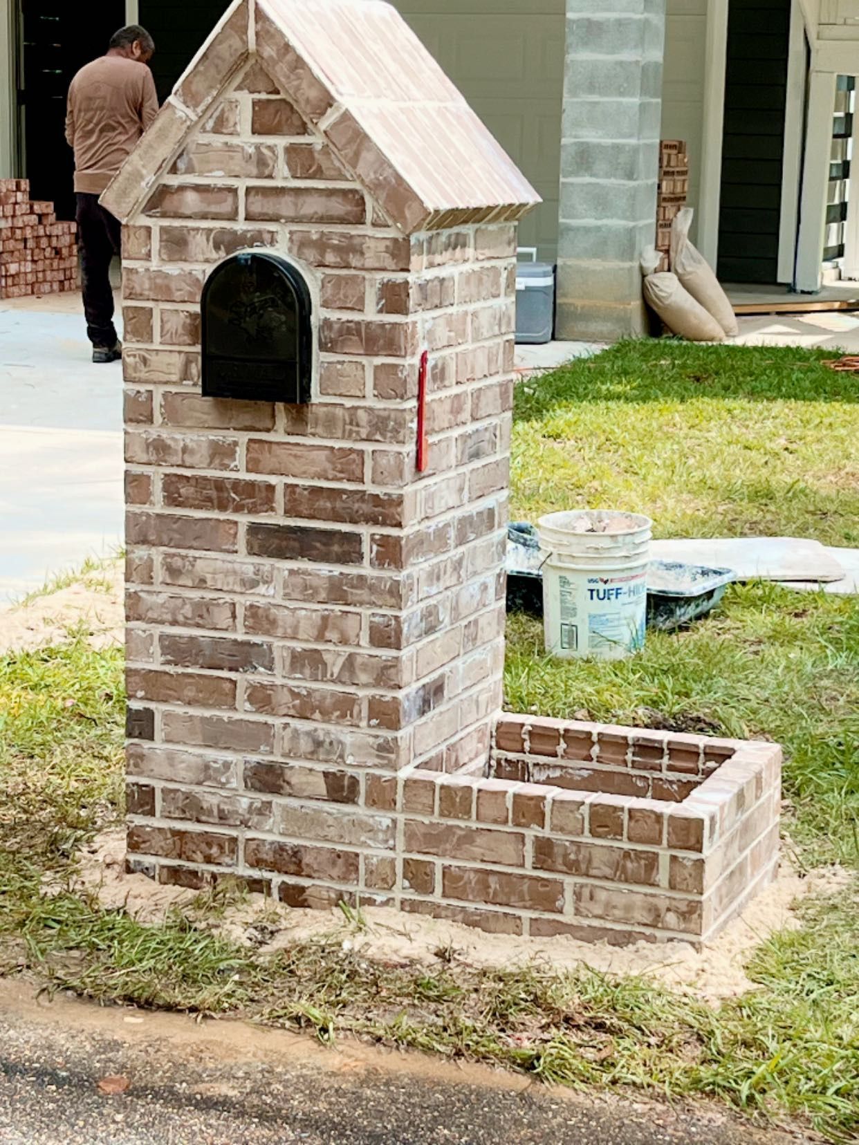 A newly built, light-brown brick mailbox structure with a peaked roof and a small attached planter on a grass lawn.