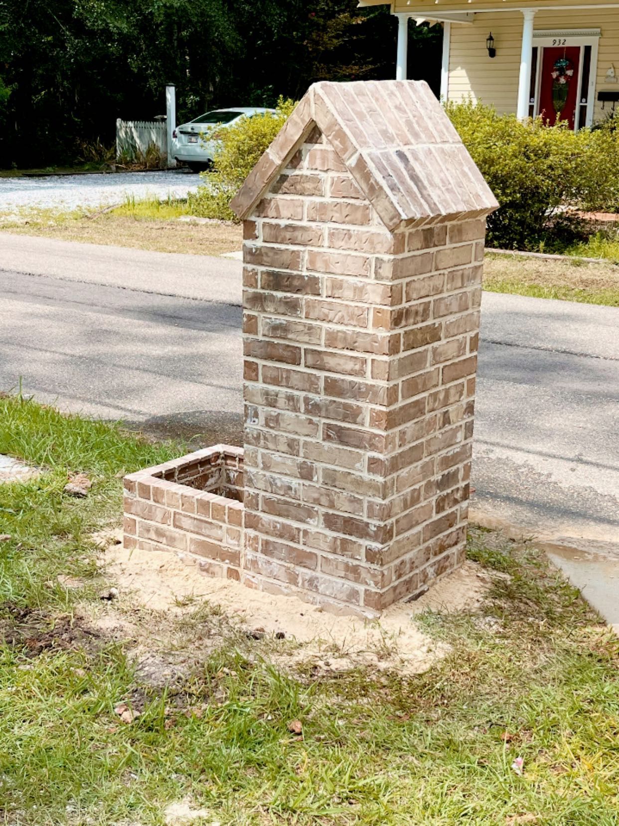 A brick mailbox structure with a peaked roof, standing on a grass lawn next to a paved road.