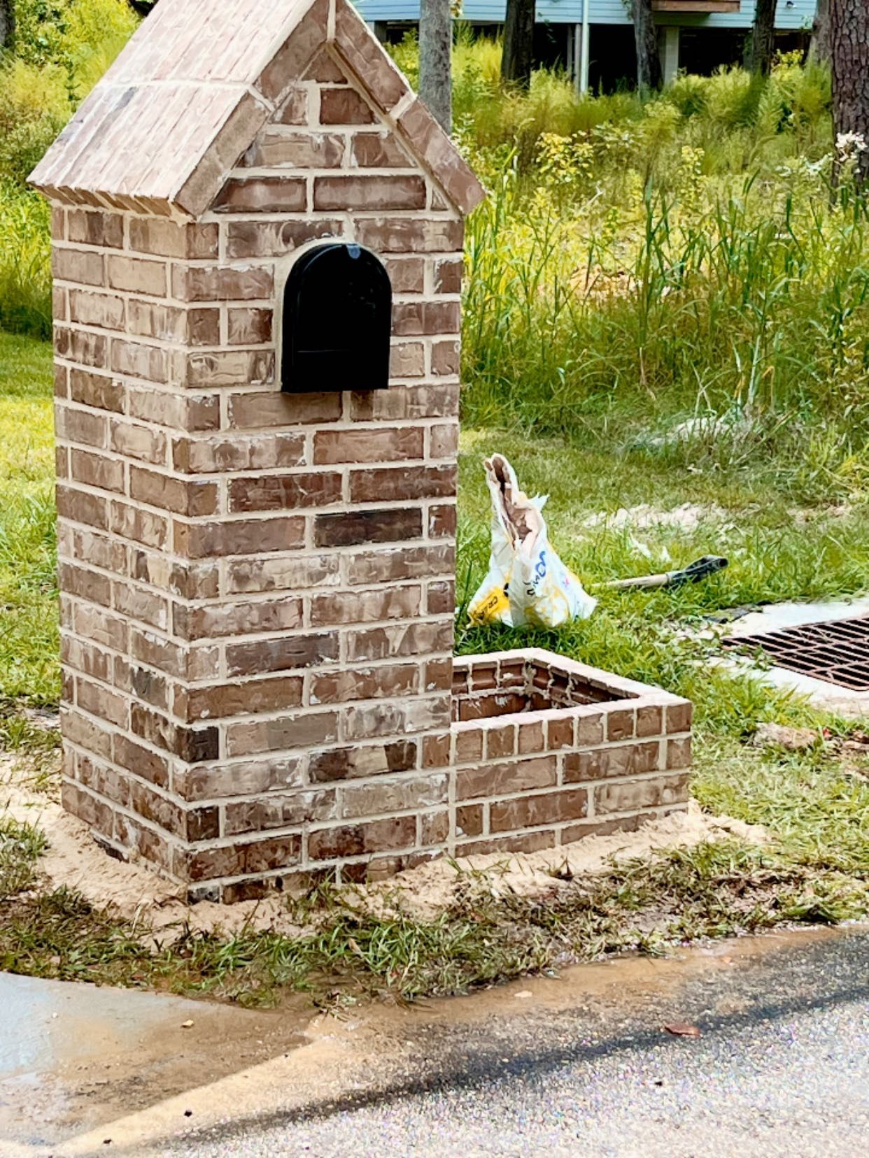 A brick mailbox structure with a black mailbox, featuring an attached planter box, standing near a road and grassy area.