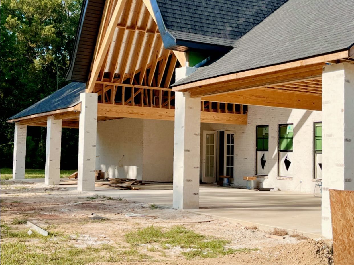 A house under construction with a white brick exterior, large support columns, and an unfinished porch area.