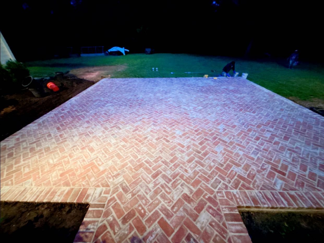Newly installed herringbone brick patio in a backyard at dusk, featuring a decorative border and surrounding dark soil.