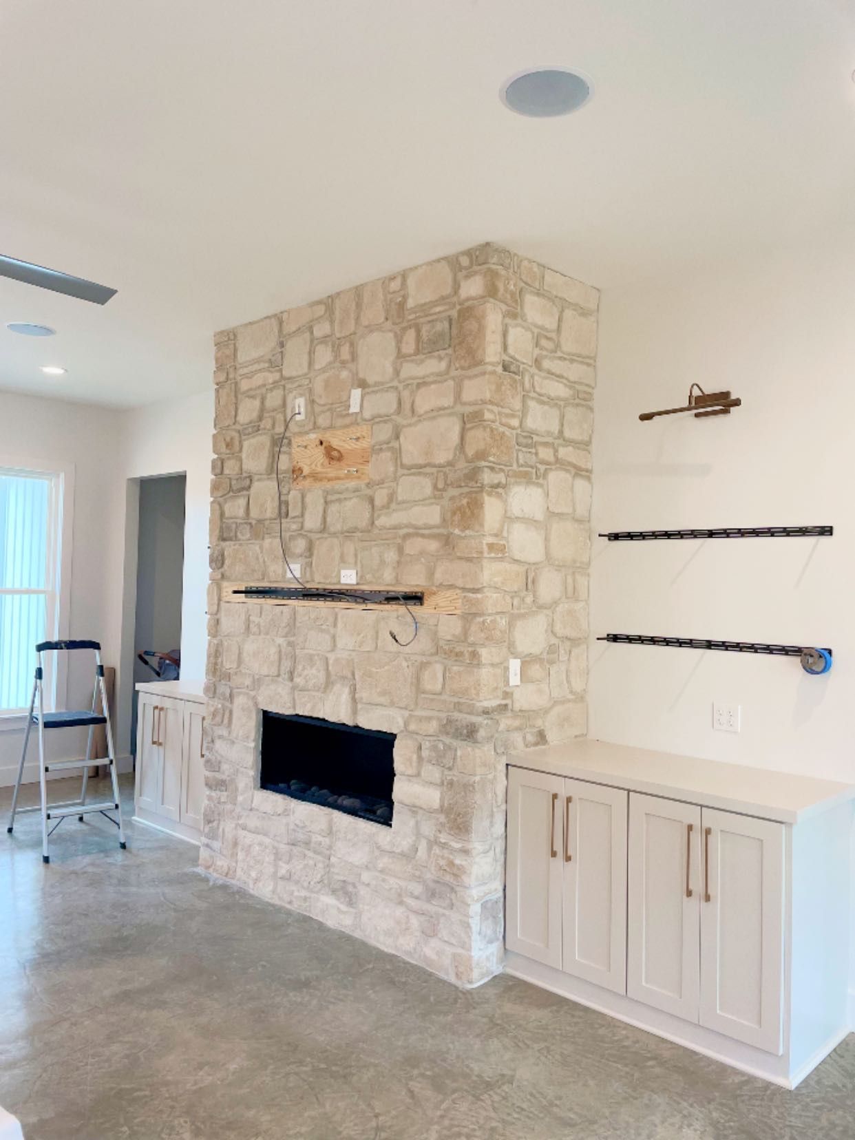 A stone fireplace sits between two white cabinets with dark shelving in a room with a concrete floor and a step ladder.