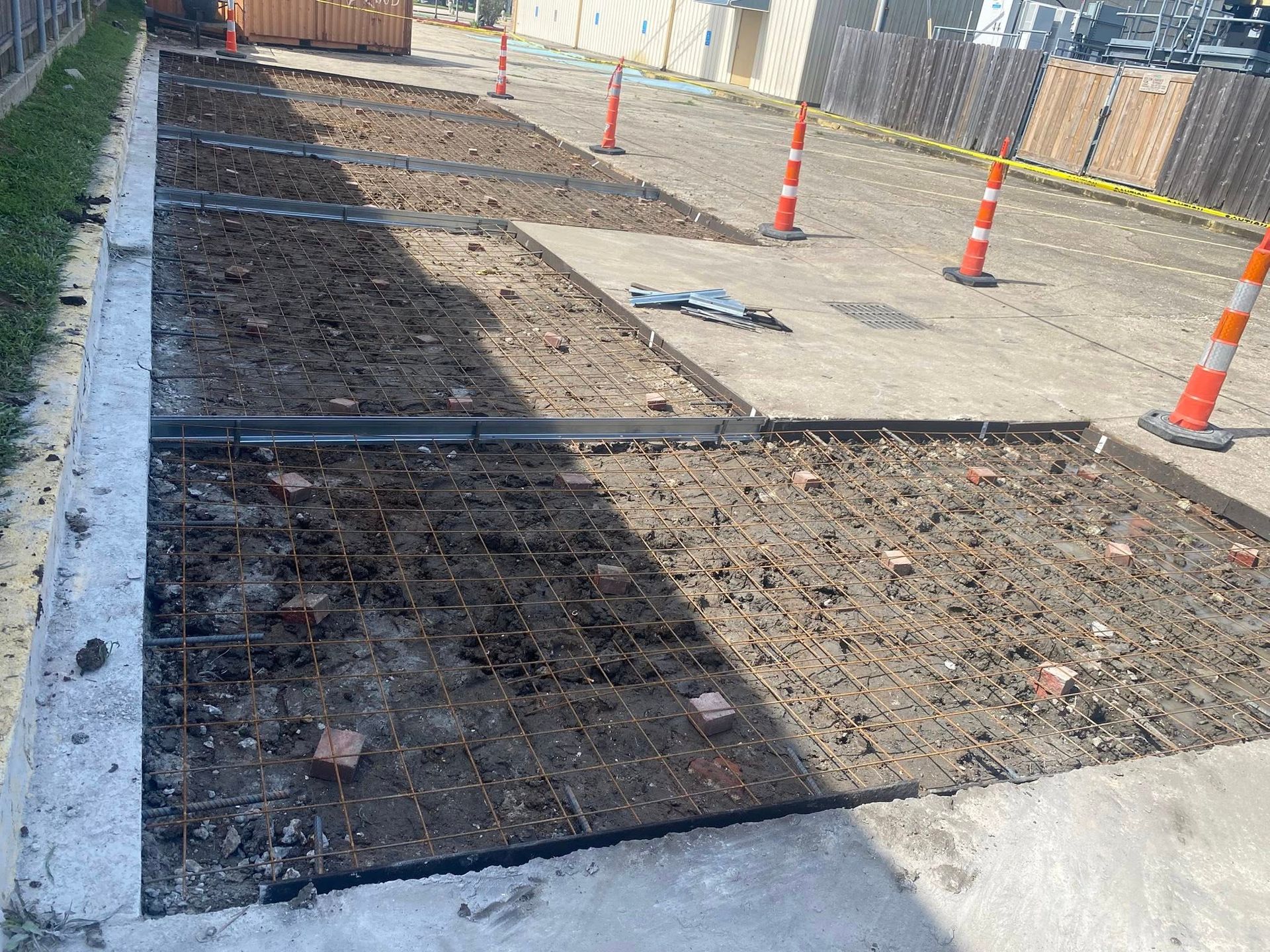 A construction site shows a rectangular area prepared for pouring concrete, featuring metal rebar mesh and orange cones.