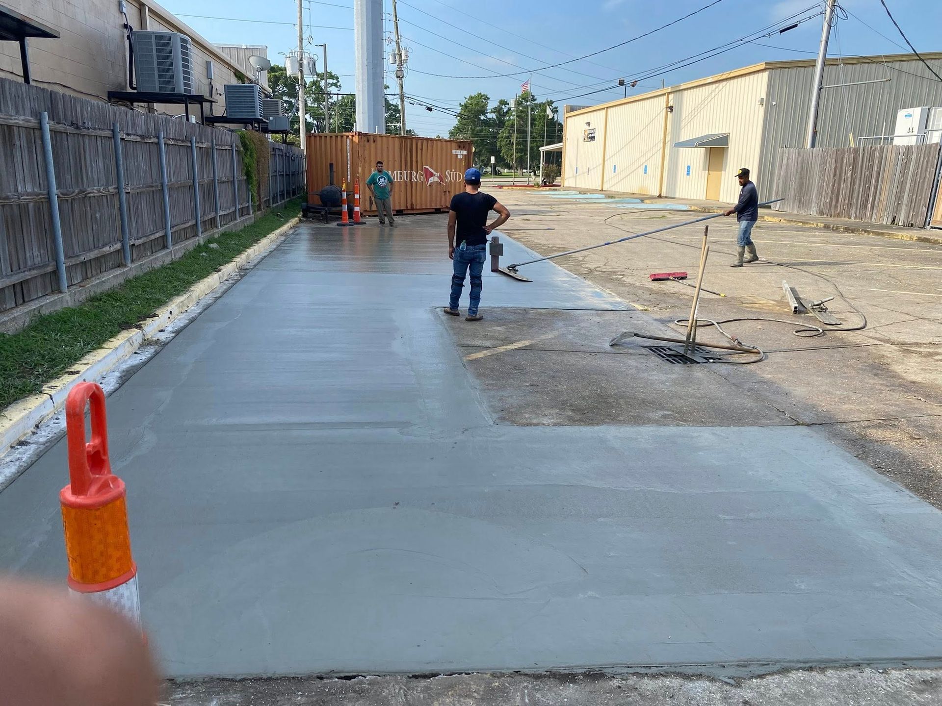 Workers stand on a newly poured concrete surface in an outdoor area beside a fenced building and a storage container.