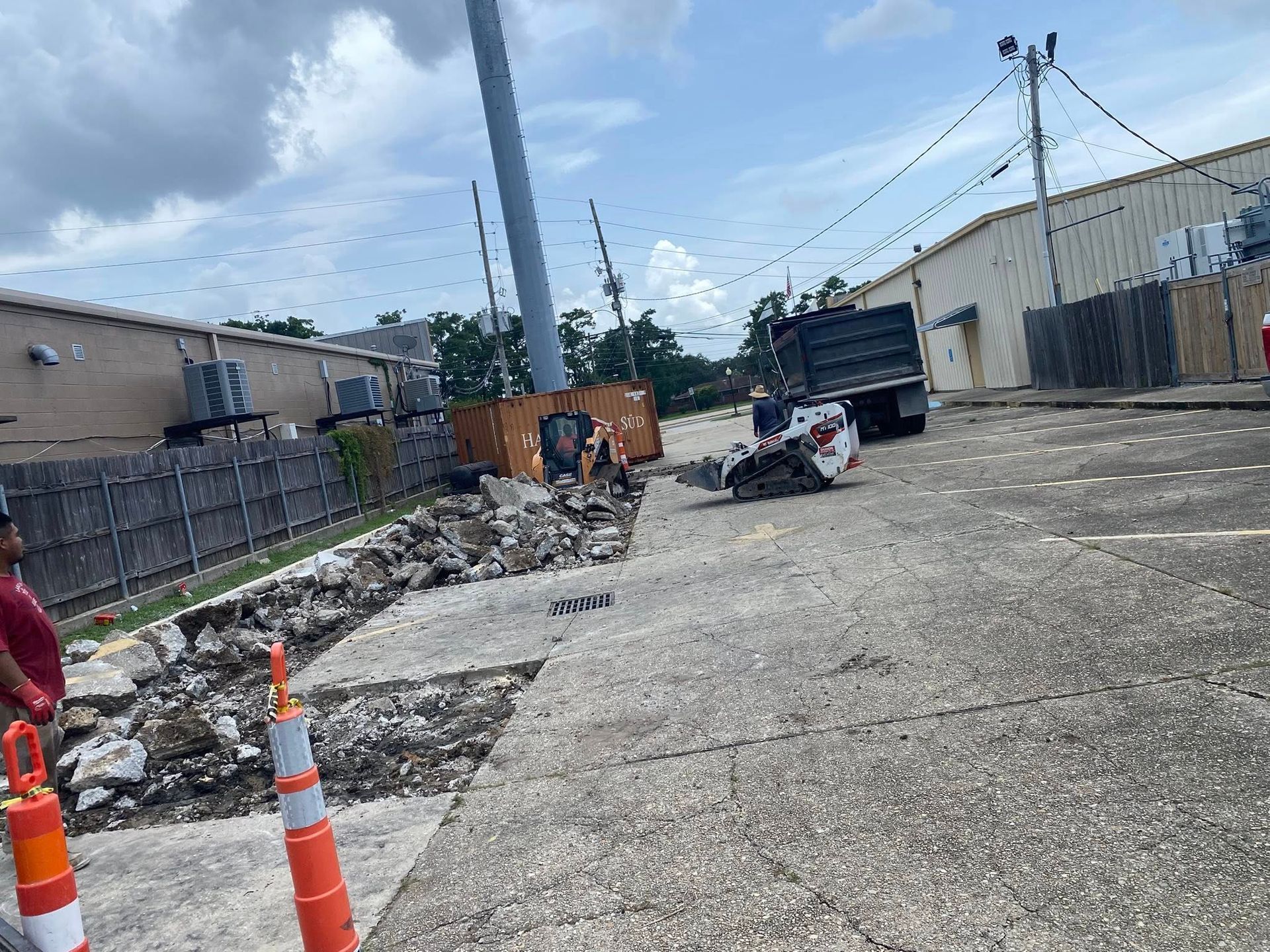 A skid steer operates in a parking lot near a fenced building, removing concrete debris into a dump truck.