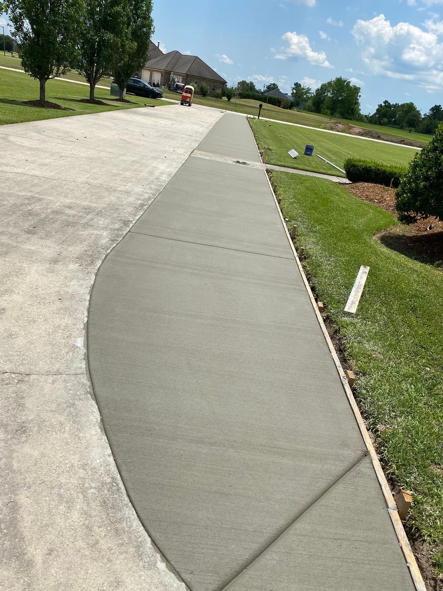 A freshly poured concrete sidewalk border along an existing driveway, set in a grassy residential landscape.