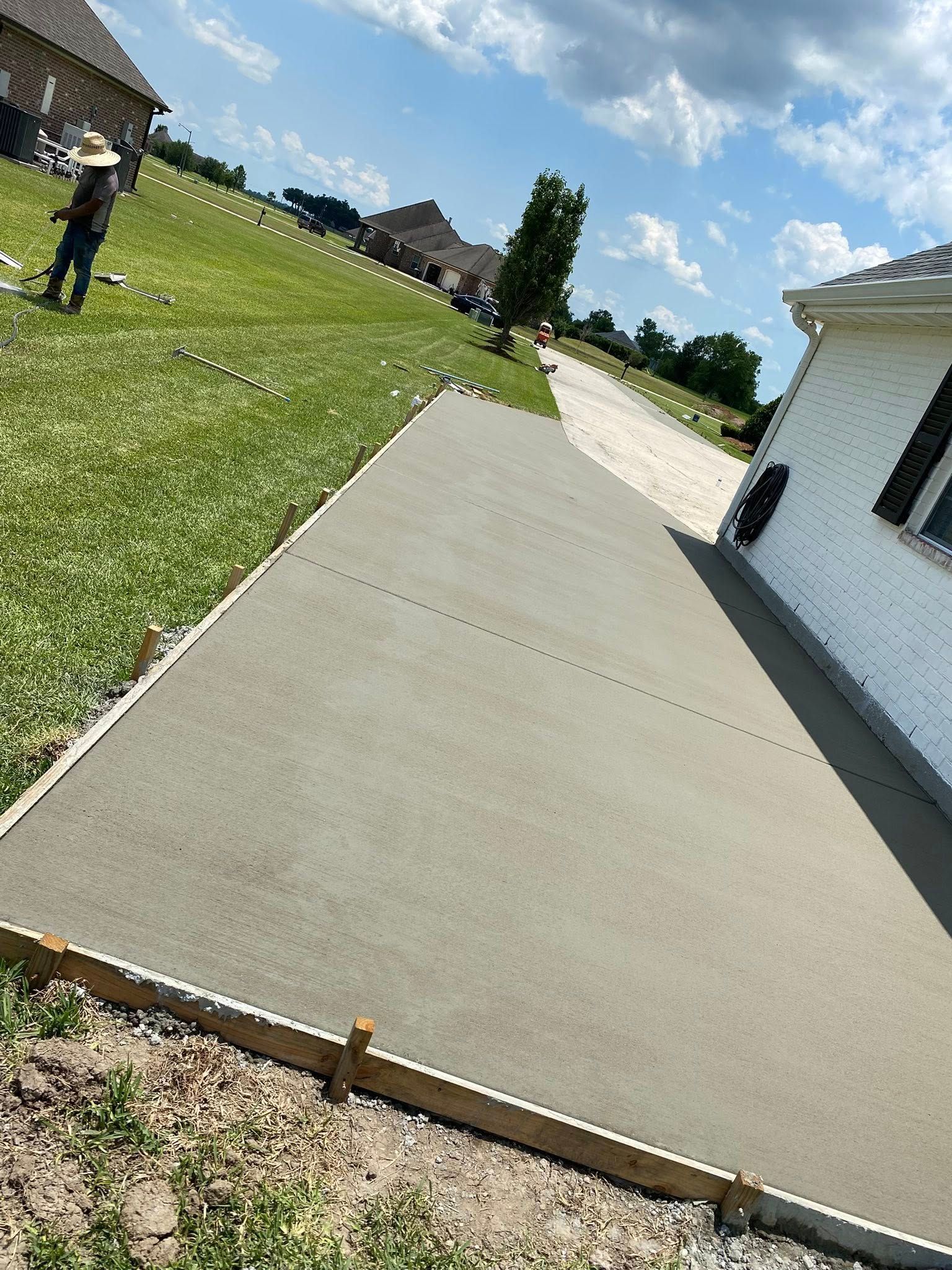 A freshly poured concrete patio slab borders a white house next to a grassy yard under a bright, sunny sky.