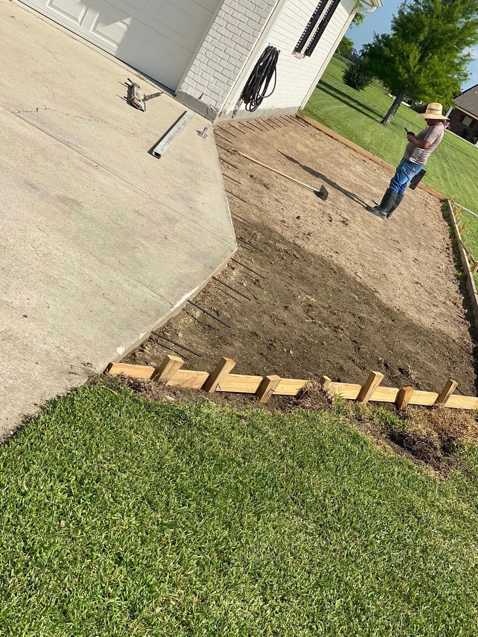 A person in a straw hat stands in a yard where wooden forms are being prepped to extend a concrete driveway.