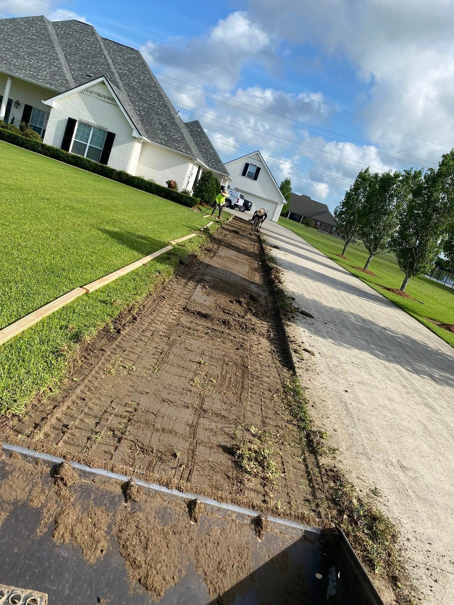 A view from machinery clearing a strip of dirt alongside a gravel driveway and green lawn between suburban houses.