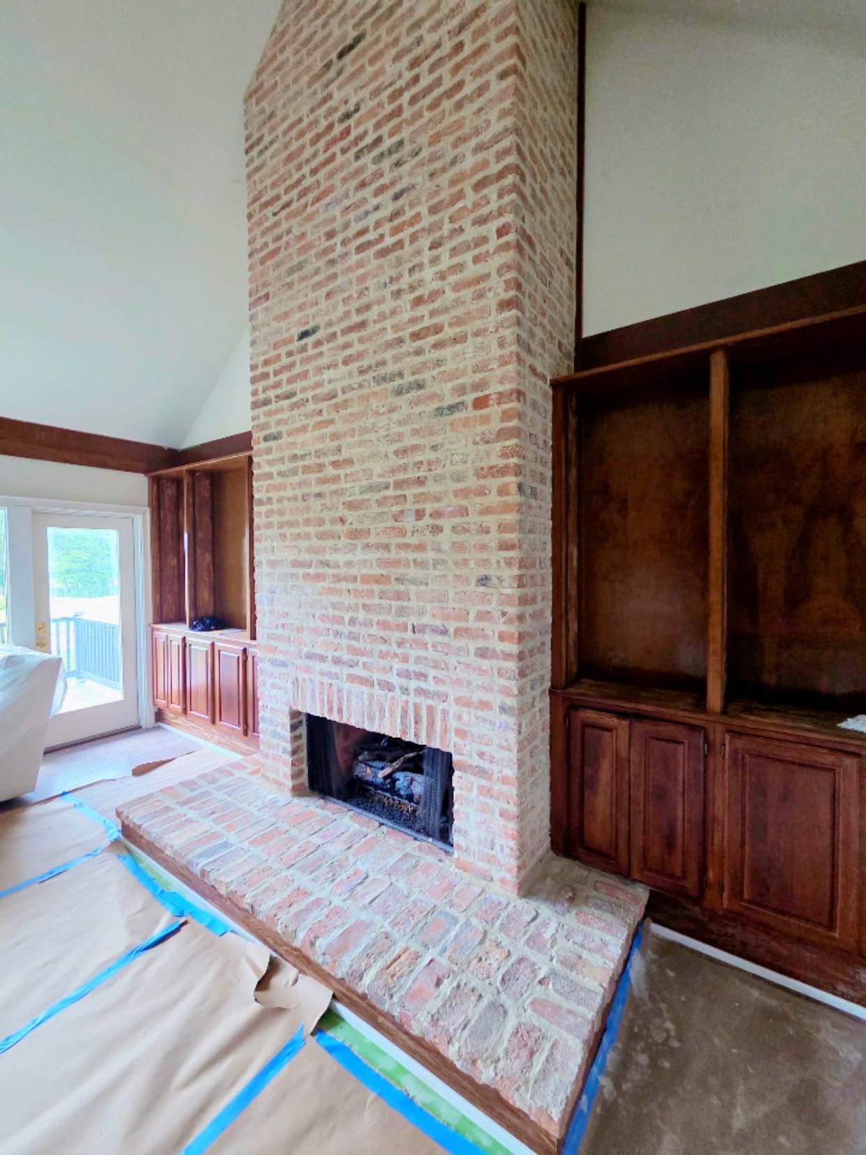 A tall, light-toned brick fireplace with a central hearth, flanked by dark wooden built-in shelving in a vaulted room.