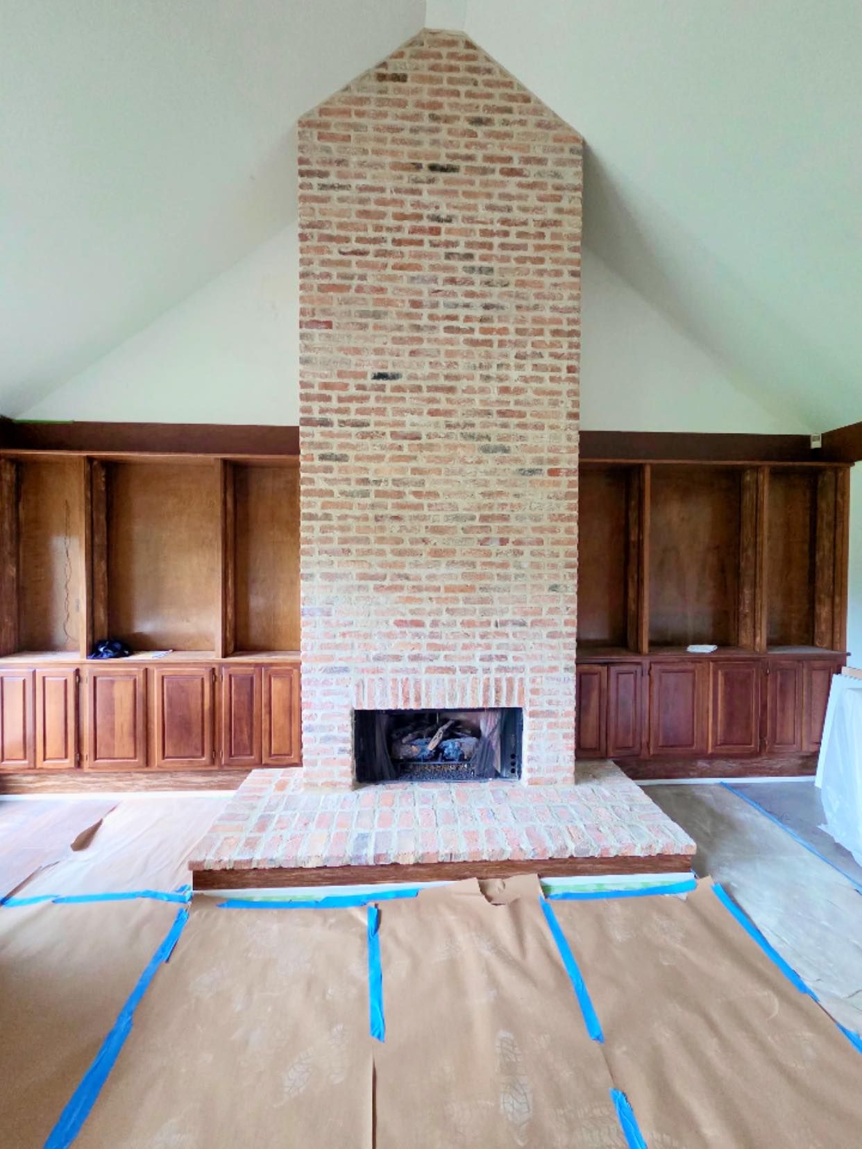 A tall brick fireplace stands centered between built-in wooden cabinets in a room with a vaulted ceiling and floor covers.