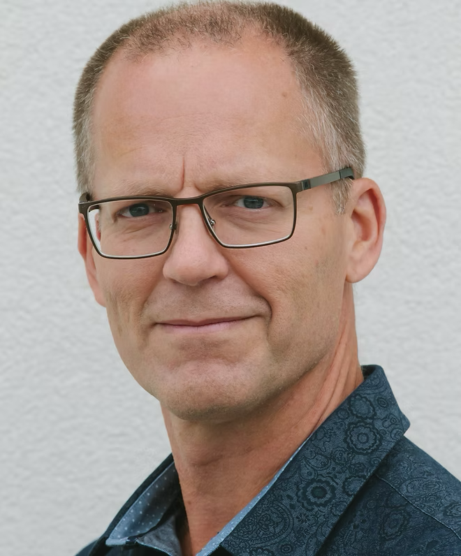 A man in a white shirt with polka dots is smiling with his arms crossed