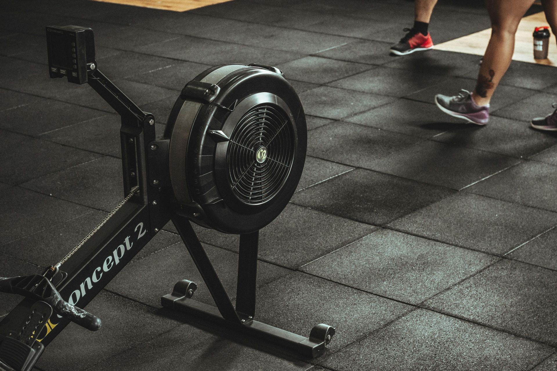 Rowing machine in a gym. People in athletic shoes are nearby on a black rubber floor.