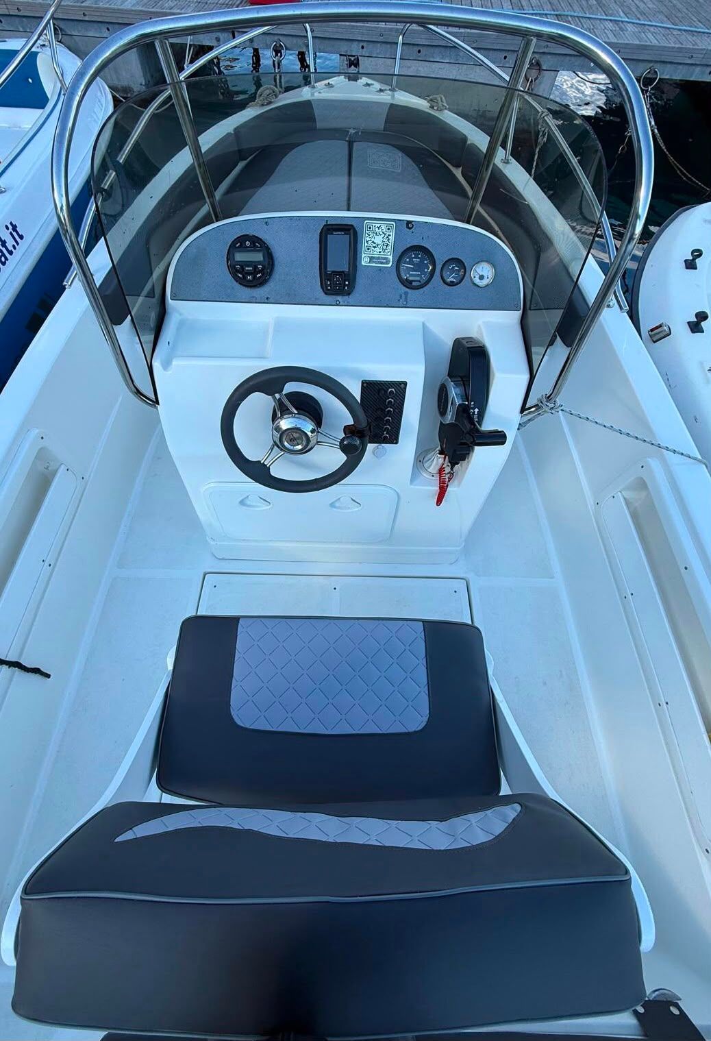 Abitacolo di una barca con cuscini e tendalino blu navy, affacciato su un porticciolo e acque calme al crepuscolo.
