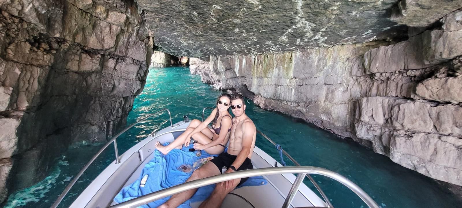 Due persone si rilassano su una barca all'interno di una grotta marina rocciosa con acqua turchese.