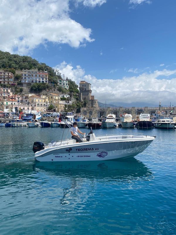Motoscafo bianco su acque blu in un porto con yacht, edifici sulle colline e un cielo nuvoloso.
