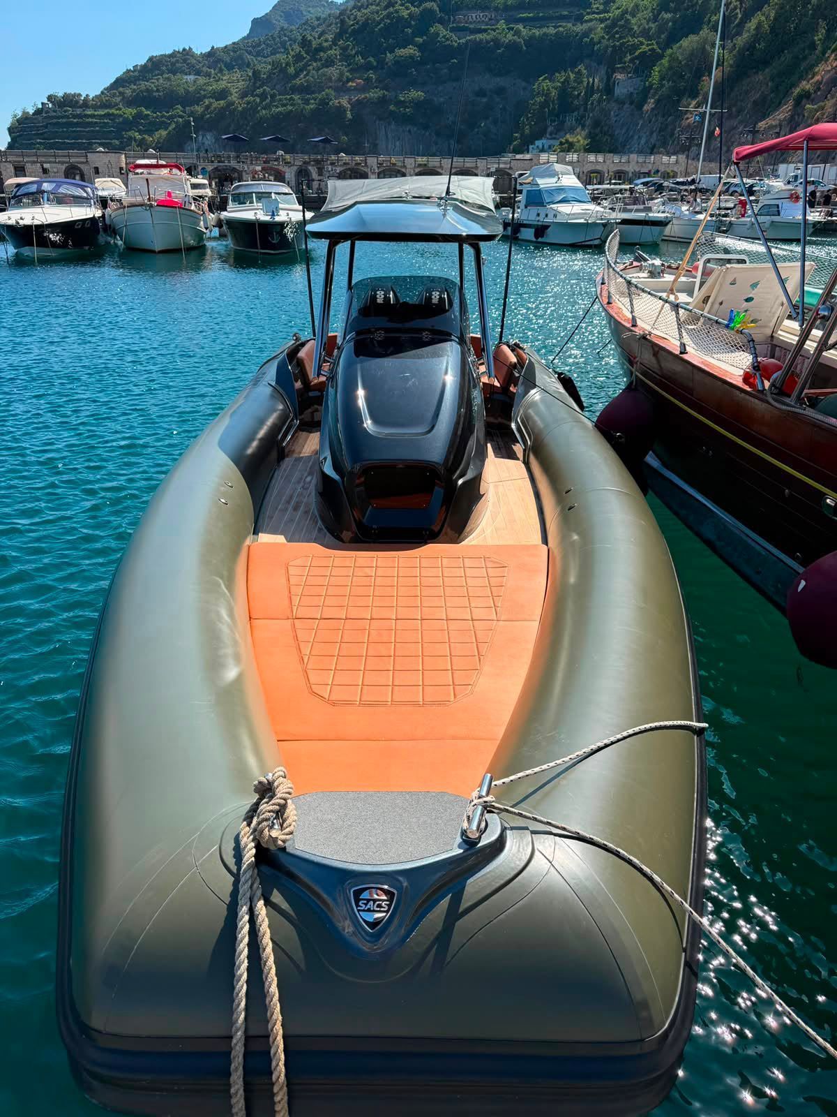 Gommone gonfiabile con ponte arancione ormeggiato in un porto turchese, con barche e costa rocciosa sullo sfondo.
