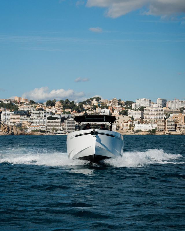 Motoscafo bianco che fende l'acqua blu con lo skyline di una città costiera sullo sfondo.