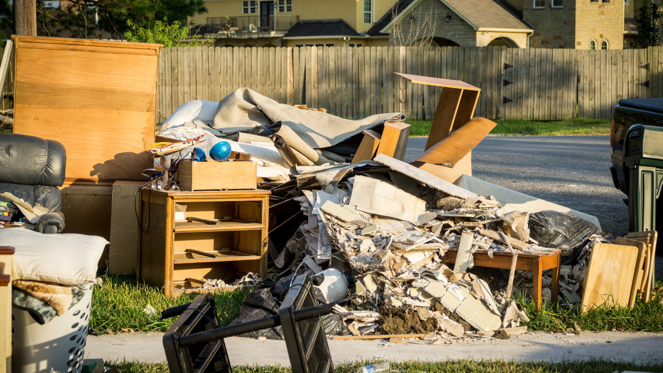 Pile of household debris on a lawn, including furniture, broken wood, and drywall, next to a street.