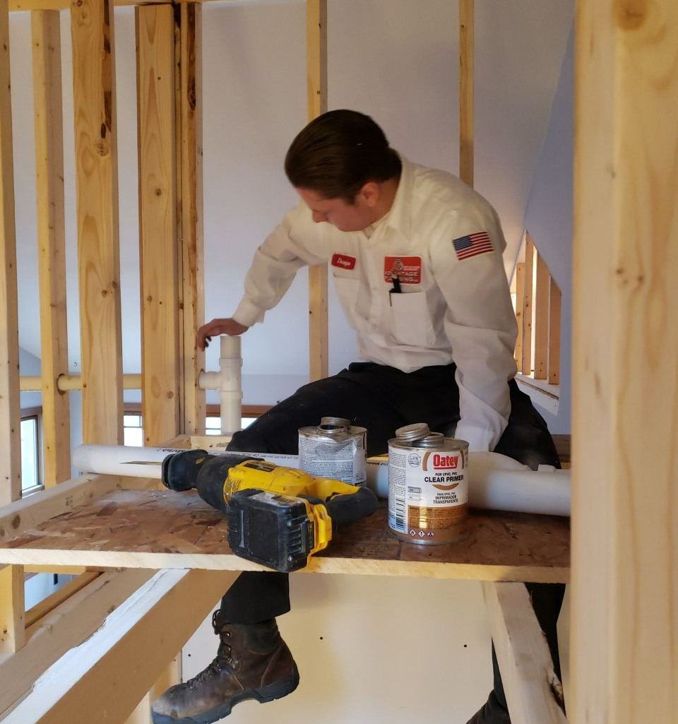 Plumber installing pipes in a framed wall; tools, glue, and a level are nearby.
