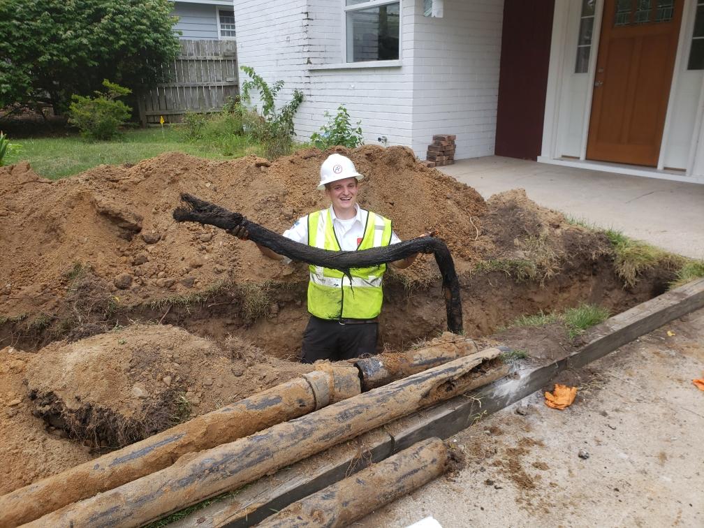 Man in safety vest holds a black pipe in a dirt trench, near a house.