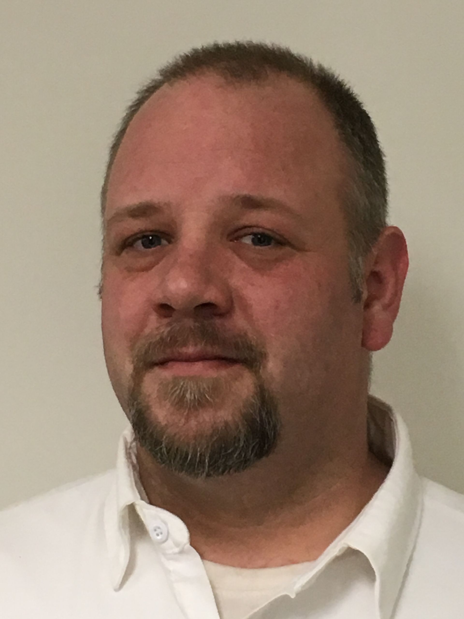 Man with short brown hair and beard, wearing a white collared shirt, smiles gently, against a plain white background.
