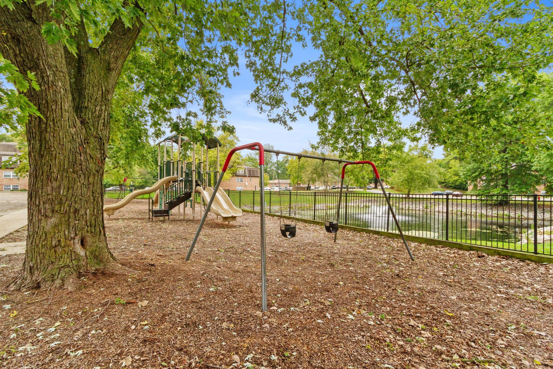 Playground with swings, slides, and tree, near a fence and water, on a sunny day.