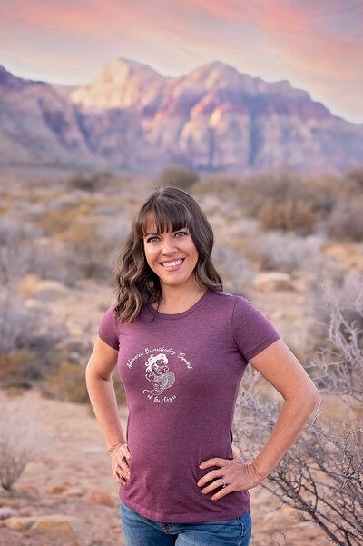 Woman in a purple shirt smiling with hands on hips in a desert landscape with mountains at sunset