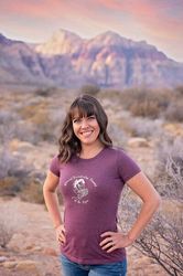 Woman in a purple shirt smiling with hands on hips in a desert landscape with mountains at sunset