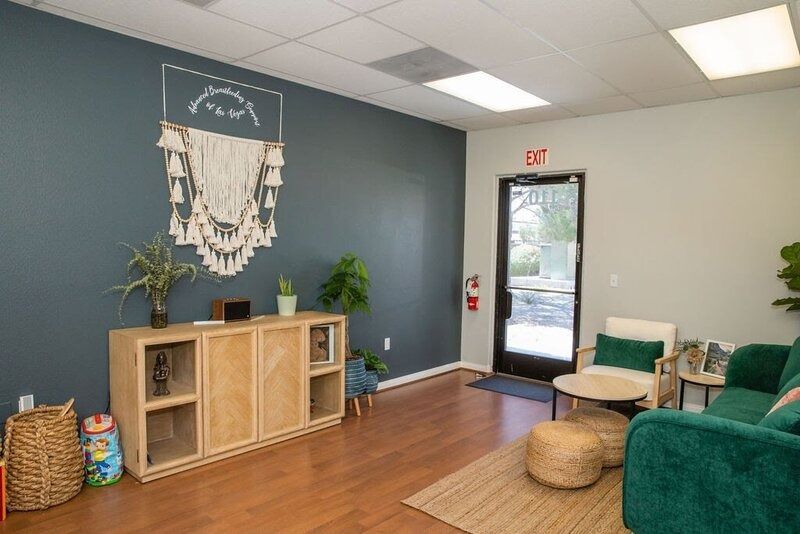 Cozy waiting area with green couch, wicker stools, wood cabinet, plants, and a glass exit door