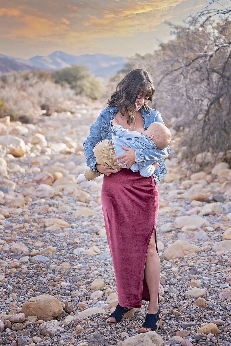 Woman holding a baby on a rocky desert trail at sunset, with mountains in the background.