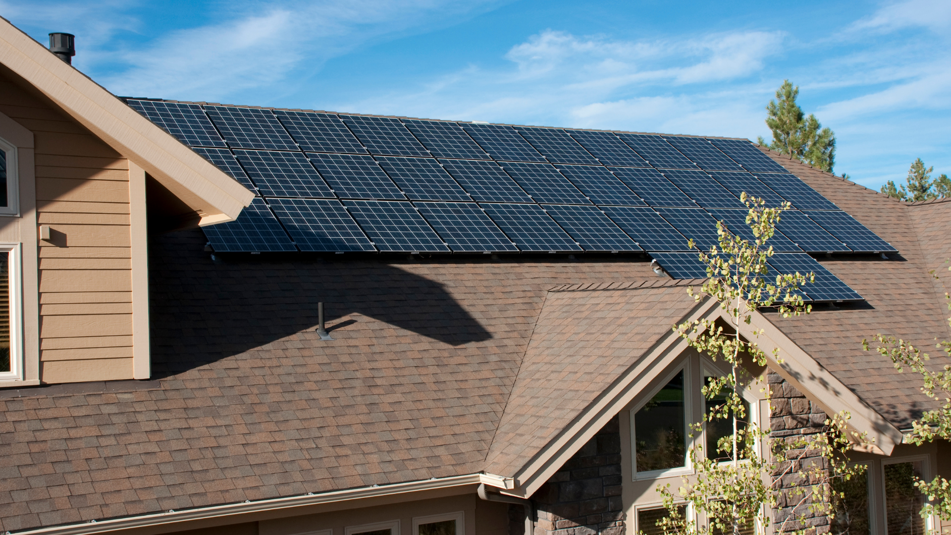 Solar panels installed across a multi-gabled brown shingle roof of a house under a clear blue sky.