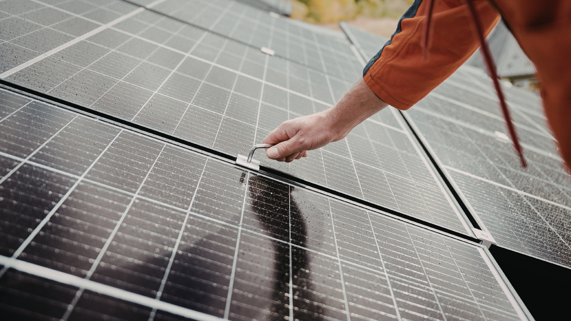 A person in an orange jacket installs a metal clamp to secure solar panels mounted on a roof.